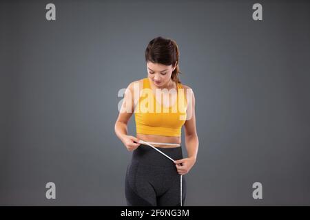 Il concetto di uno stile di vita sano. Donna atletica snella in abbigliamento sportivo davanti a una parete grigia che misura la vita con un nastro. Una ragazza giovane con Foto Stock