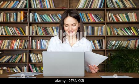 Donna giovane sorridente in blusa bianca elegante lavora con carte vicino a un computer portatile contemporaneo a un tavolo contro grandi scaffali della libreria piena di libro Foto Stock