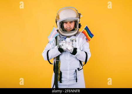 Cosmonaut gay transessuale maschile con gesto serio in tuta spaziale e casco, tenendo lgtbi bandiera arcobaleno e bandiera transgender, su sfondo giallo. Foto Stock