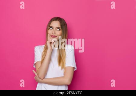 Carino giovane donna in camicia whte sta tenendo la mano sul mento, guardando il lato e pensando. Girata in casa su sfondo rosa. Foto Stock