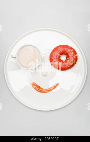 vista dall'alto della faccia sorridente fatta di latte in tazza, ciambella, zucchero cubetto e marmellata su vassoio rotondo su bianco Foto Stock