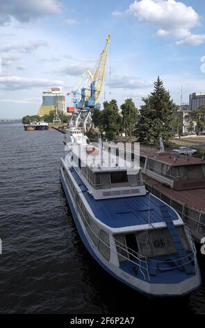 Barche da crociera sul fiume ormeggiate accanto al molo, con gru e un blocco di appartamenti oltre, fiume Dnieper, Dnipro, Ucraina. Foto Stock