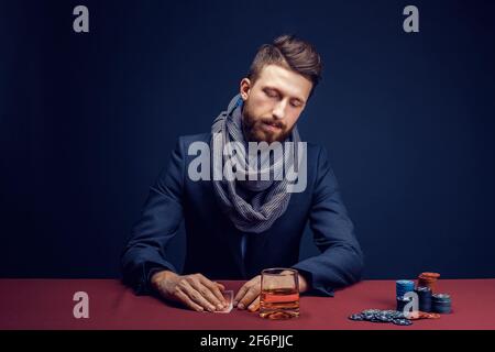 Elegante uomo bearded che gioca nel casinò scuro, fumare sigaro, bere whiskey Foto Stock