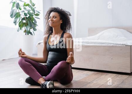 Sorridente donna afro-americana incinta seduta in yoga posa in camera da letto Foto Stock