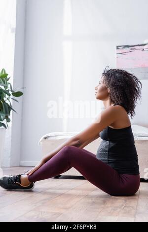 Vista laterale di una donna sportiva afro-americana incinta seduta sul pavimento in camera da letto Foto Stock