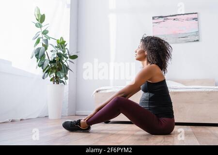 Vista laterale di una donna sportiva afro-americana incinta sorridente sul pavimento in camera da letto Foto Stock