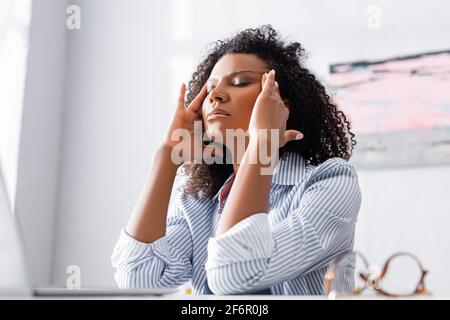 Donna afro-americana stanca che soffre di mal di testa vicino al computer portatile e. occhiali in primo piano sfocato Foto Stock