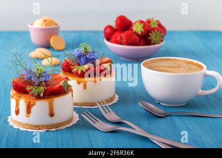 Due mini cheesecake alla fragola rotondi senza cottura con caramello al sale e metà di fragole, impreziosite da veri fiori blu. Tavolo di legno blu. Foto Stock