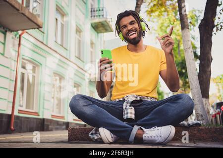 Ritratto fotografico di un ragazzo seduto a terra che ascolta la musica con il cellulare sorridente sulla strada della città in estate Foto Stock