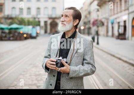 Uomo anziano in piedi sulla strada della città con fotocamera retrò sul collo. Turista maschile in abiti eleganti godendo di splendida architettura della città vecchia. Foto Stock