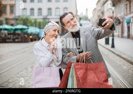 Coppia anziana allegra che prende selfie sullo smartphone moderno mentre si alza in strada con borse per la spesa in mani. Concetto di famiglia, tecnologia e acquisto. Foto Stock