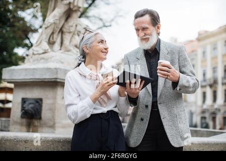 Sorridente coppia anziano che usa il tablet digitale per strada. Famiglia matura in abiti eleganti seduti vicino alla vecchia fontana e bere caffè. Trascorrere il tempo insieme. Foto Stock