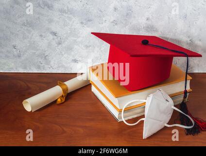 Ancora vita con un cappello rosso di laurea su alcuni libri accanto ad un diploma e una maschera su una scrivania di legno. Concetto di laurea e di distanziamento sociale Foto Stock