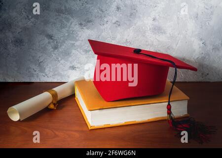 Ancora vita con un cappello rosso di laurea su un libro accanto ad un diploma su una scrivania di legno. Concetto di studi e di laurea Foto Stock