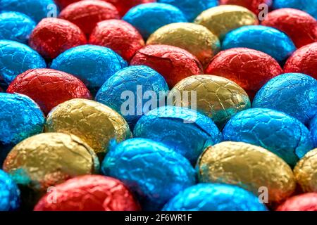 Primo piano di molte uova di pasqua al cioccolato avvolte in lamina blu, rossa e dorata. Sfondo pasquale con fuoco selettivo. Foto Stock