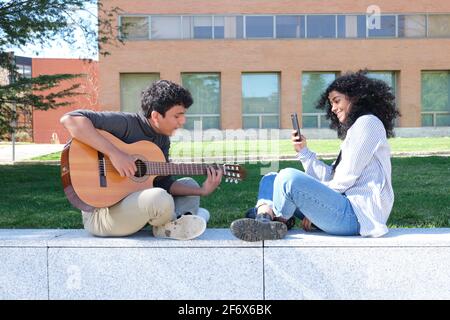 Giovane latino che suona la chitarra e il suo amico che lo registra con lo smartphone nel campus universitario. Vita universitaria, generazione millenaria. Foto Stock