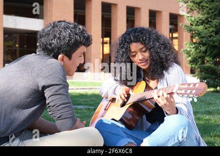 Giovane latino che insegna a suonare la chitarra ad una giovane donna latina nel campus universitario. Vita universitaria, generazione millenaria. Foto Stock