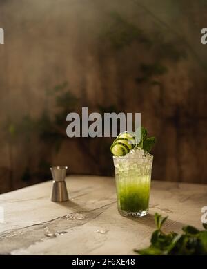 Ancora vita con un bicchiere di limonata vegetale fredda con cetriolo, menta e ghiaccio tritato. Bevanda rinfrescante, jigger di metallo ed erbe fresche in luce naturale. Foto Stock