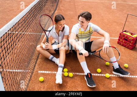 Coppia allegra seduta sul pavimento del campo da tennis con racchette nelle mani Foto Stock