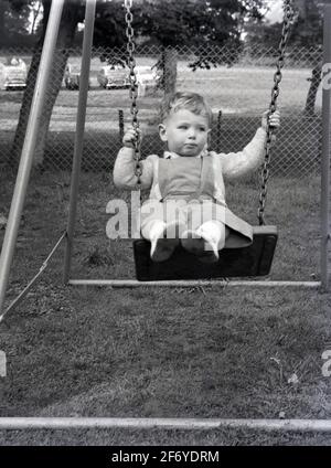 1956, storico, fuori in un parco giochi, da solo, un ragazzino seduto su una piccola altalena che tiene sulle sue catene metalliche con un'altalena. Un'attività preferita per i bambini di tutte le età, l'oscillazione è infatti molto befica per i bambini piccoli aiutandoli a sviluppare i sottili pattini del motore di forza di presa, di mano, di braccio e di coordinazione delle dita. Sviluppa anche i muscoli di un bambino e sviluppa il suo senso di equilibrio. Foto Stock