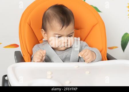 Primo piano di un bambino di sei mesi seduto su una sedia alta utilizzando la pinza afferrare per prendere uno spuntino dalla sedia alta vassoio Foto Stock