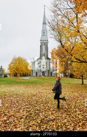 Giovane bionda, indossa un cappello mentre si gioca nelle foglie autunnali con una vecchia chiesa cattolica sullo sfondo in una giornata piovosa. Foto Stock
