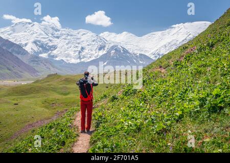 Un giovane escursionista maschile con uno zaino in azione di Fotografare la bellezza panoramica del picco di Lenin innevato (montagne di Pamir) nel campo base Foto Stock