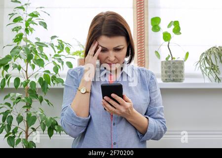 Triste donna di mezza età con telefono a casa vicino a. finestra Foto Stock