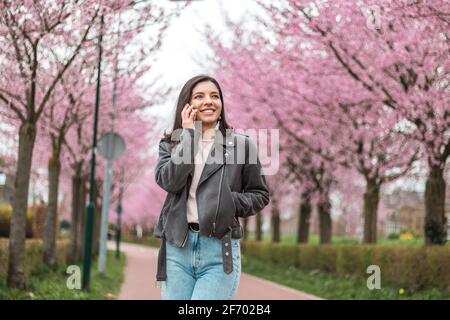 giovane donna allegra e fiduciosa di etnia mista telefona con amici o parenti amati che camminano da soli in fiore parco con alberi rosa Foto Stock