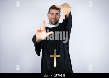 Giovane uomo ispanico che indossa uniforme sacerdote in piedi su sfondo bianco sorridente rendendo cornice con mani e dita con volto felice Foto Stock