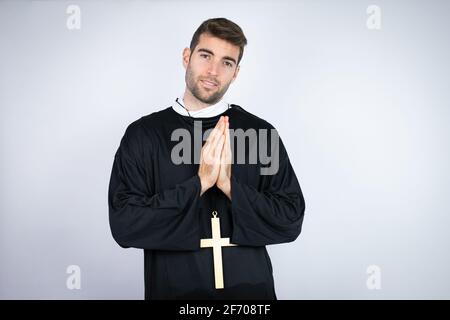 Giovane uomo ispanico che indossa uniforme sacerdote in piedi su sfondo bianco pregando con le mani insieme faccia allegra e felice. Foto Stock