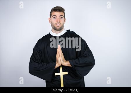 Giovane uomo ispanico che indossa uniforme sacerdote in piedi su sfondo bianco pregare e pregare con le mani insieme con l'espressione della speranza faccia molto emo Foto Stock