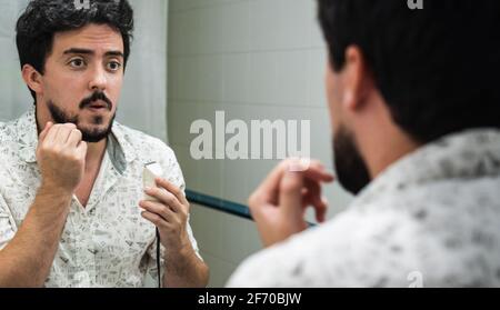 giovane con barba che rifiniva la barba con un regolabarba, rifinendo di fronte a uno specchio in bagno Foto Stock