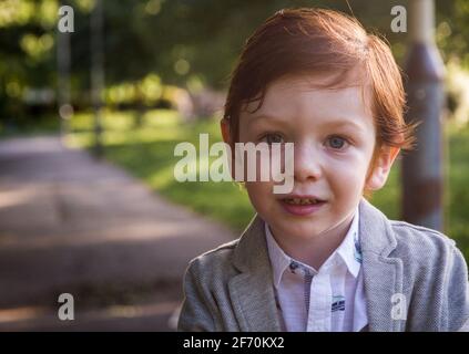 Ritratto di un simpatico ragazzo rosso dagli occhi blu che indossa un blazer grigio e una camicia bianca in un parco in una giornata di sole Foto Stock