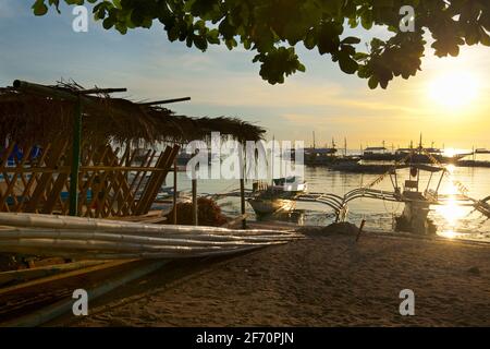 La spiaggia sabbiosa di Logon, isola di Malapascua, con le canoe Outrigger sulla spiaggia al tramonto. Mare di Visayan, Cebu, Filippine. Foto Stock