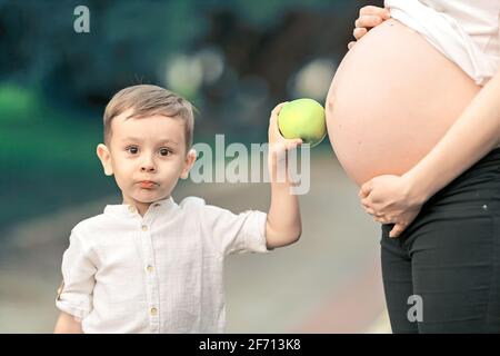 Ritratto di un ragazzino accanto ad una madre incinta in una giornata estiva nel parco. Il bambino tiene una mela vicino al pancia grande della madre. Cura e amore in famiglia. Foto Stock