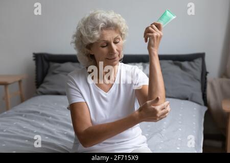 Donna anziana matura anni 60 che applica crema sul gomito in camera da letto Foto Stock