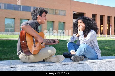 Giovane coppia latina innamorata che suona la chitarra e canta nel campus universitario. Vita universitaria, generazione millenaria. Foto Stock