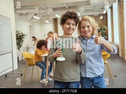 La nostra invenzione. Ritratto di due ragazzi felici che mostravano i pollici mentre tenevano il giocattolo meccanico che assemblavano sulle classi di artigianato. Bambini che posano per la macchina fotografica a. Foto Stock