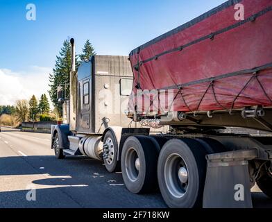 Trattore semi-camion classico americano a lunga percorrenza con cofano di banco trasporto fisso con imbragature coperte con carico commerciale pesante su letto piatto semi Foto Stock