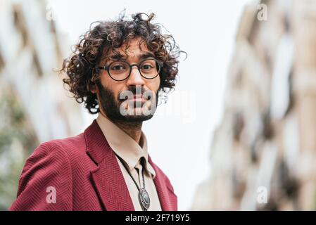ritratto orizzontale del giovane caucasico sulla strada guardando la macchina fotografica. Indossa abiti e occhiali casual alla moda. Ha capelli ricci e una barba Foto Stock