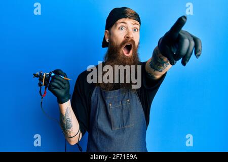 Uomo rosso con tatuaggio per barba lunga artista che indossa uniforme professionale e guanti rivolti con il dito sorpreso davanti, bocca aperta stupito espressione, Foto Stock