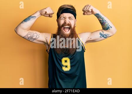 Uomo rosso con barba lunga che indossa uniforme da basket mostrando muscoli sorridenti e ridendo forte perché divertente scherzo pazzo. Foto Stock