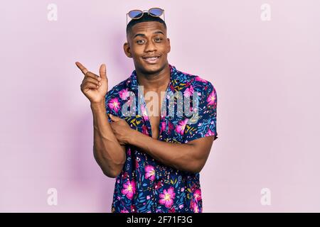 Giovane uomo nero che indossa una camicia hawaiana e occhiali da sole con un grande sorriso sul viso, puntando con la mano e il dito a lato guardando la fotocamera. Foto Stock