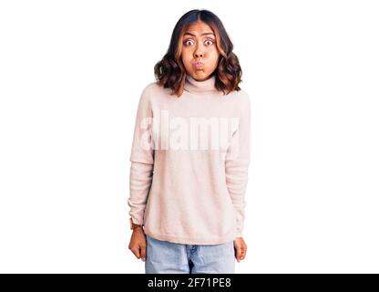 Giovane bella donna mista da corsa con maglione invernale a turleneck che soffia guance con viso divertente. Bocca gonfia con aria, espressione pazza. Foto Stock