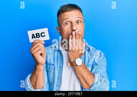 Bell'uomo muscolare che tiene lettere abc su carta che copre la bocca con la mano, scioccato e impaurito per errore. Sorpresa espressione Foto Stock