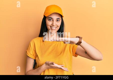 Giovane donna ispanica che indossa la divisa di consegna e cappuccio gesturing con le mani che mostrano grande e grande segno di misura, simbolo di misura. Sorridente guardando la camma Foto Stock