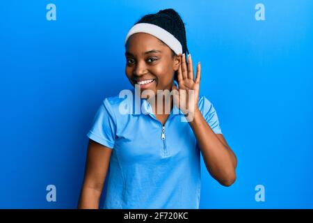 Donna afroamericana con capelli intrecciati che indossa un giocatore di tennis uniforme sorridente con mano sopra l'orecchio ascolto e l'udito di voce o pettegolezzi. Sordità c Foto Stock