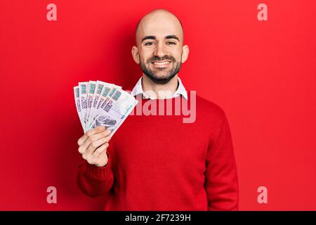 Giovane uomo calvo che tiene le banconote egiziane delle libbre che osservano positive e. felice in piedi e sorridente con un sorriso sicuro che mostra i denti Foto Stock