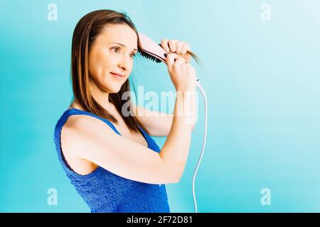 Bella giovane donna spazzolando i suoi capelli con ioni spazzola elettrica soffio asciugacapelli con fondo blu. Concetto di cura dei capelli Foto Stock